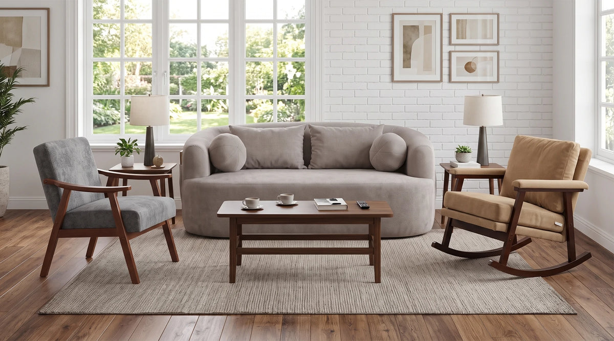 A staged living room scene featuring a light gray upholstered loveseat centered on a woven area rug, with a wooden coffee table in front, a gray accent chair on the left, and a tan rocking chair on the right. Large white-framed windows, white brick wall, and framed art are visible in the background.